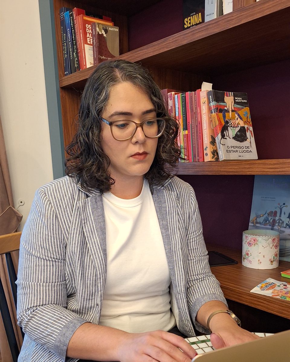 Karoline Pereira, de óculos e blazer listrado, trabalhando no notebook em frente a uma estante de livros